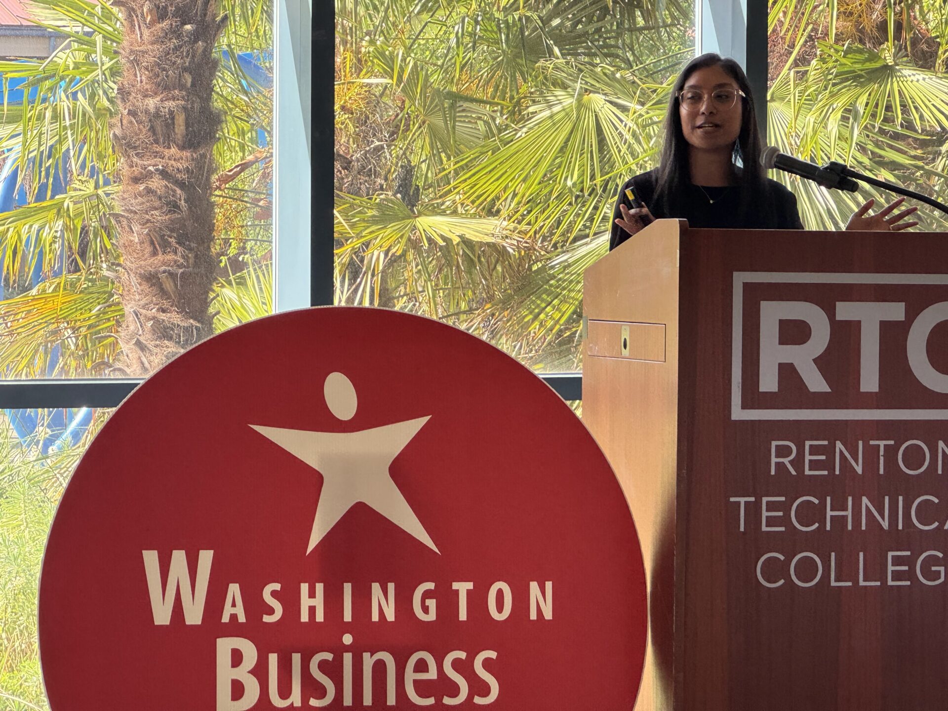 A woman stands behind a podium reading "Renton Technical College" a cutout of the Washington Business Week logo is in the foreground.