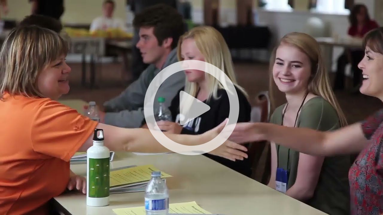 A woman shakes the hands of several students. A play button is overlayed.